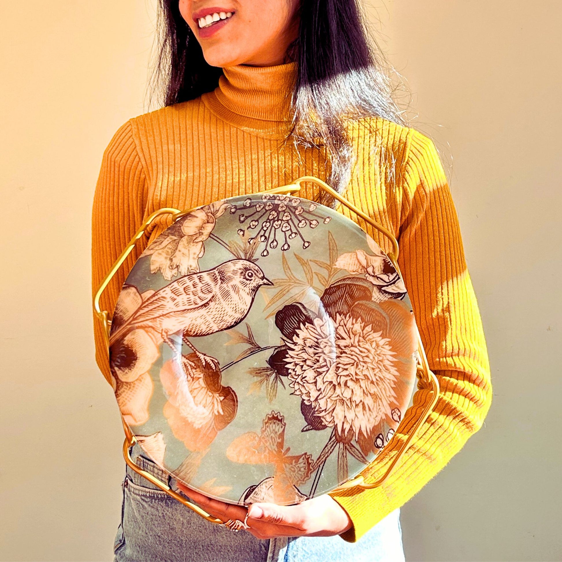 Model holding a Cake Stand with Bird and flower print on a blue backdrop. The stand has an interlinked chain design in brushed gold.