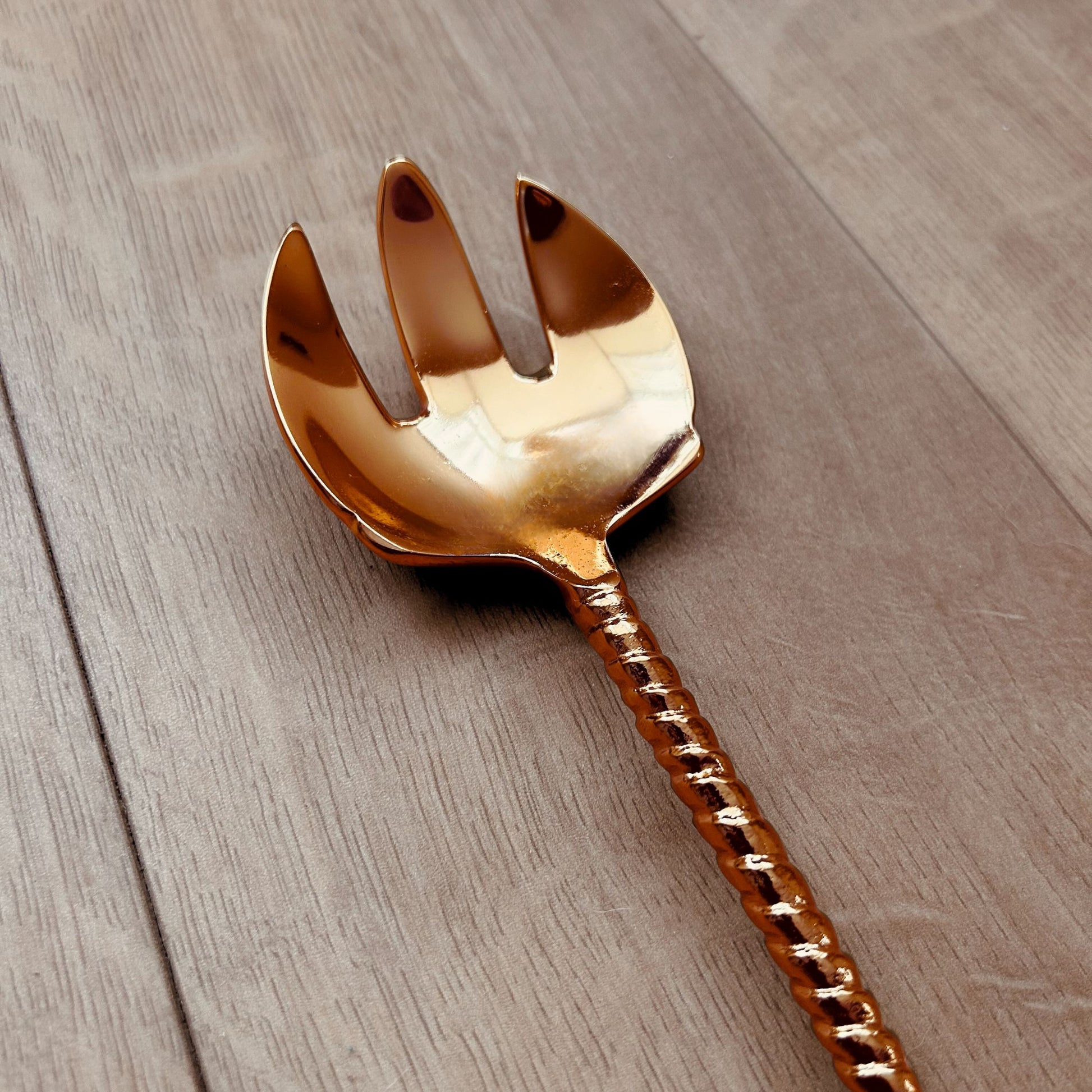 Close Up of a Gold Finish Stainless Steel Salad Serving Fork with a twine and leaf handle. It is kept on a wooden backdrop.