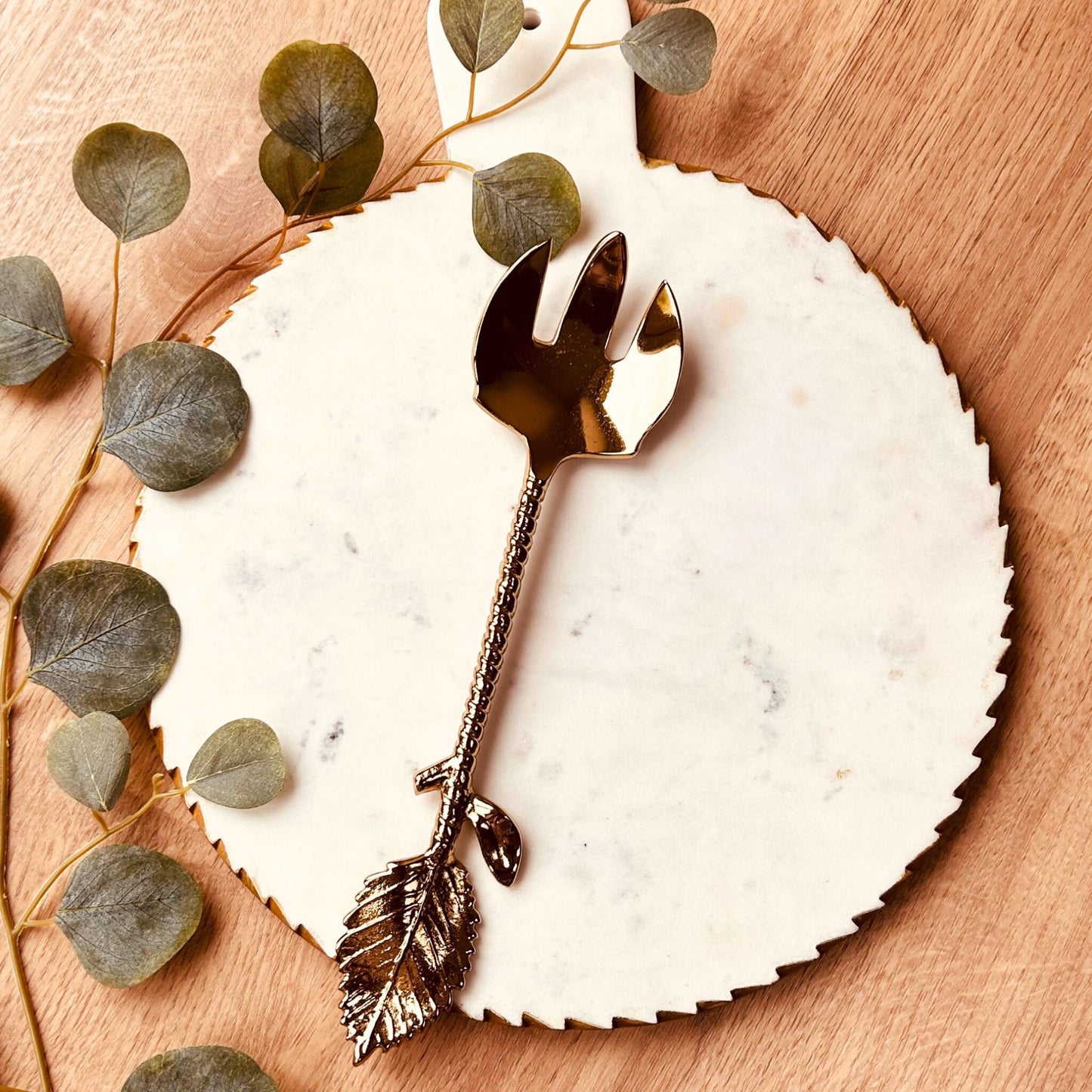 Gold Finish Stainless Steel Salad Serving Fork with a twine and leaf handle. It is kept on a white marble serving board with leaves on the side.