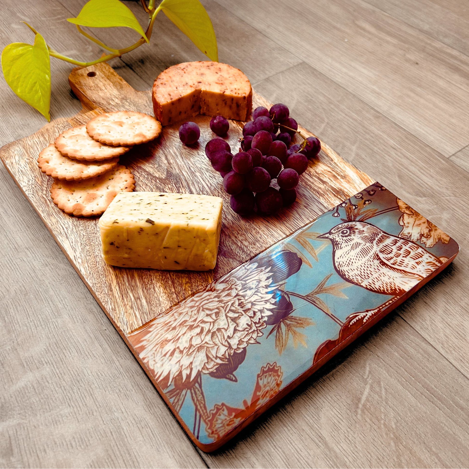 Mango Wood Charcuterie Board with handle with crackers, cheese and grapes. The Charcuterie Board has a nature, bird and flowers print at the bottom of the board.