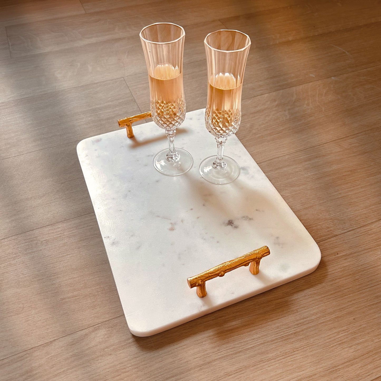 Rectangular White Marble Tray with Metal handles in gold finish kept on a wooden surface. It has two champagne glasses kept on it.