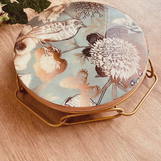 Cake Stand with Bird and flower print on a blue backdrop. The stand has an interlinked chain design in brushed gold.