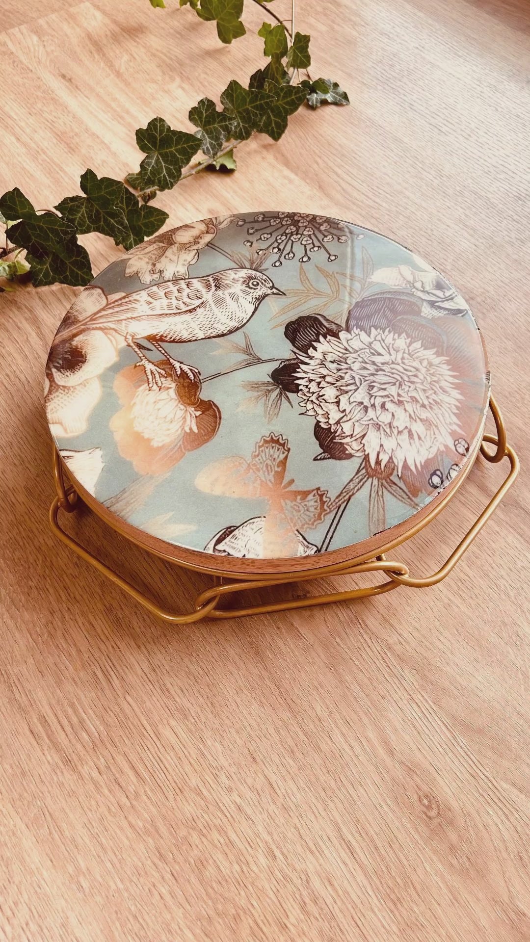 Cake Stand with Bird and flower print on a blue backdrop. The stand has an interlinked chain design in brushed gold.